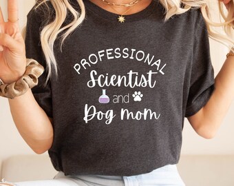 Könnte beinhalten: Ein dunkelgraues T-Shirt mit dem Text "Professional Scientist and Dog Mom" in Weiß. Neben dem Text befinden sich ein violetter Becher und ein Pfotenabdruck.