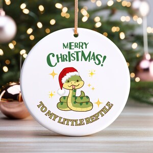 May include: White ceramic Christmas ornament with a green snake wearing a red Santa hat. The ornament says "Merry Christmas! To My Little Reptile".