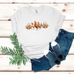 May include: White t-shirt with a graphic of three chickens and strawberries. The chickens are brown and white and the strawberries are red.