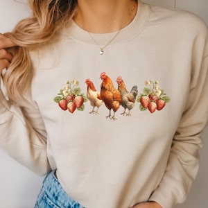 May include: A beige sweatshirt with a graphic of three chickens and strawberries. The chickens are brown and white, and the strawberries are red.