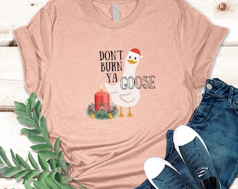 May include: A pink t-shirt with a white goose wearing a Santa hat and holding a lit candle. The text on the shirt reads "Don't burn ya goose".