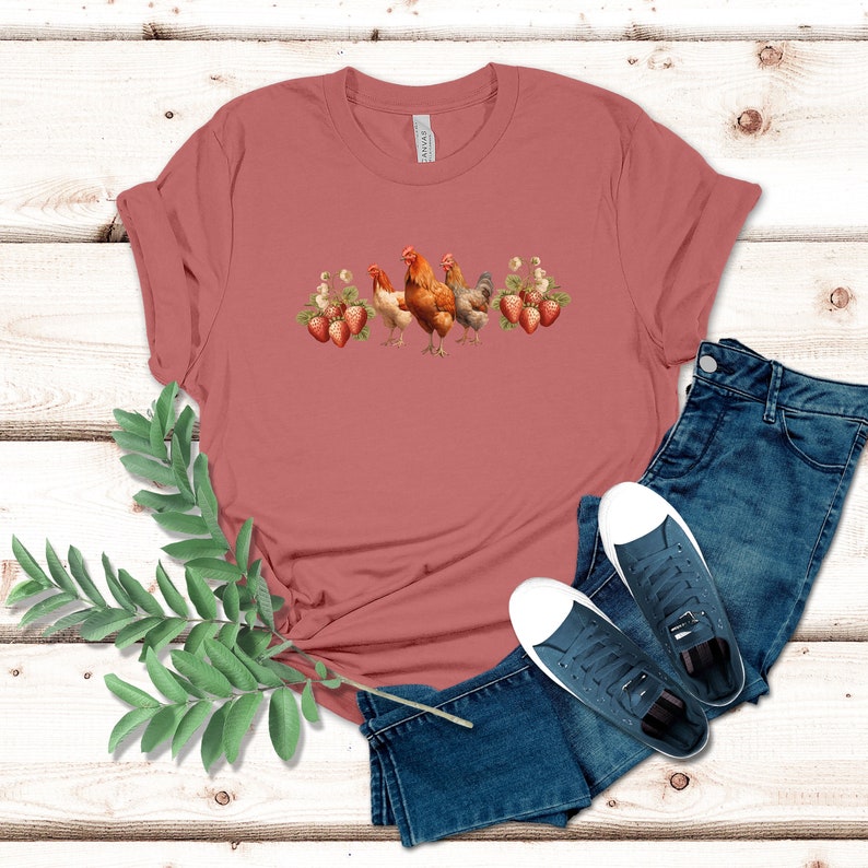 May include: A maroon t-shirt with a graphic of chickens and strawberries. The t-shirt is laid out on a wooden surface with a pair of blue jeans and white sneakers.