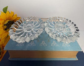 May include: Two clear glass candle holders with a flower-like design. The candle holders are sitting on a blue book with gold-colored pages. A yellow sunflower is visible in the background.