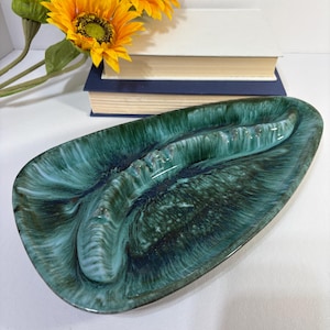 May include: An elongated, teardrop-shaped ceramic ashtray featuring a glossy glaze in shades of deep forest green, teal, and black. The surface displays a swirling, marbled pattern with a raised, textured central ridge containing multiple notches.