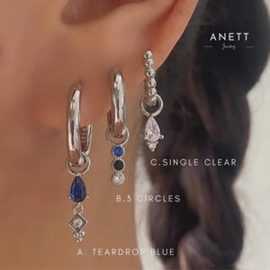 May include: Silver hoop earrings with hanging charms. One earring has a teardrop blue gem and a diamond-shaped charm. Another has three circles, and the third has a single clear teardrop gem. The text "ANETT Jewelry" is in the upper right.