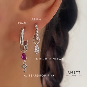 May include: Two silver hoop earrings with charms. The earring on the left has a pink teardrop charm with a small diamond charm below it. The earring on the right has a clear teardrop charm with a small diamond charm below it. The text "A. TEARDROP PINK" is below the left earring and the text "B. SINGLE CLEAR" is below the right earring. The text "ANETT Jewellery" is at the bottom of the image.