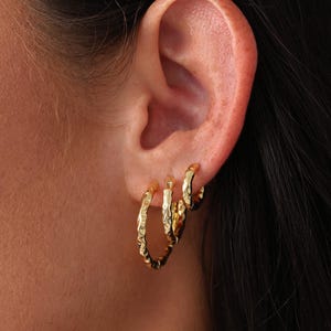 May include: Three gold hoop earrings with a hammered texture are worn on an ear. The earrings are different sizes, with the largest hoop in the front and the smallest in the back. The earrings are made of a shiny gold metal.