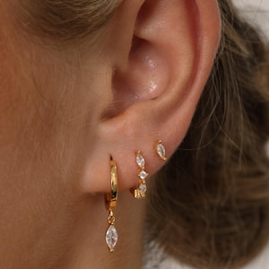 May include: Close-up of a person's ear adorned with three sets of gold earrings. The earrings include small studs, hoops with dangling teardrop-shaped gems, and hoops with multiple clear gems. The earrings are set against the backdrop of the ear and hair.