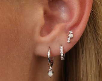May include: A silver hoop earring with a dangling opal charm, a small silver hoop earring with clear stones, and a silver stud earring featuring a central opal stone flanked by two clear stones, all worn in a single earlobe and cartilage.