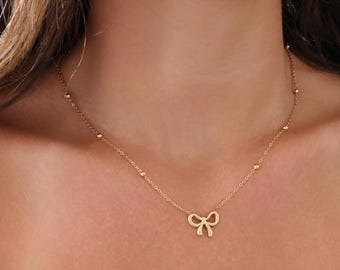May include: A gold necklace with a delicate bow pendant and small beads. The necklace is on a model's neck.