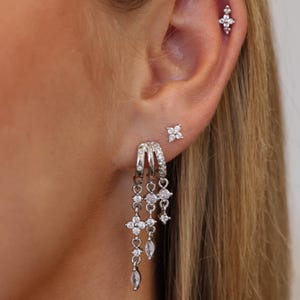 May include: A set of silver-toned earrings featuring three small hoops with dangling chains adorned with clear cubic zirconia stones in floral and marquise shapes, plus two additional small floral-shaped stud earrings on the earlobe and cartilage.