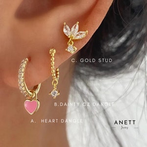 May include: Three gold earrings featuring clear cubic zirconia stones. The first earring is a hoop with a pink heart charm. The second earring is a delicate chain with a single stone charm. The third earring is a small stud with a flower design.