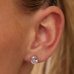 May include: A single oval-shaped stud earring featuring a clear, faceted gemstone set in a metallic prong setting. The earring is positioned on the earlobe, showcasing the reflective, brilliant cut of the stone and the secure metal base.