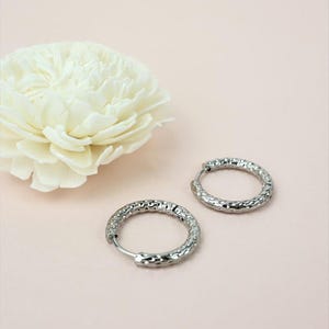May include: A pair of silver hoop earrings with a textured, hammered design. The earrings are displayed on a light pink surface, with a white flower in the background.