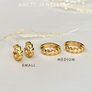 May include: Three gold hoop earrings with a heart design. The earrings are shown in three sizes: small, medium, and large. The earrings are made of gold metal and have a shiny finish.