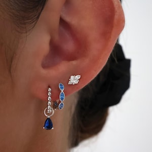 May include: Silver hoop earrings with blue gemstone accents. The earrings feature a teardrop-shaped gemstone dangling from the hoop.