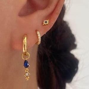 May include: Gold hoop earrings with a dangling blue gemstone and a small gold star stud earring with a black gemstone.