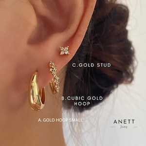 May include: An image showing three gold earrings on an ear. The earrings are a small gold hoop, a cubic gold hoop with small stones, and a gold stud with four small stones. The text "ANETT Jewelry" is in the bottom right corner.