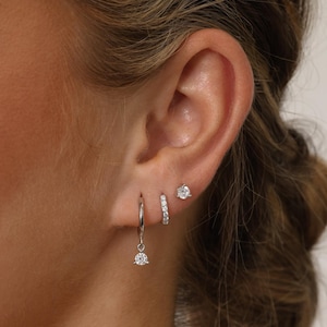 May include: An ear featuring three silver earrings. There's a small stud, a small hoop with tiny stones, and a dangling earring with a single stone. The earrings are all set with clear, sparkling stones. The earrings are a classic and elegant style.
