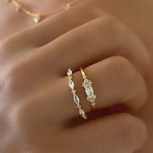 May include: Two gold rings with clear, sparkling stones. The rings are stacked on a finger. The top ring has a larger, oval-shaped stone surrounded by smaller stones. The bottom ring has a row of smaller, round stones.