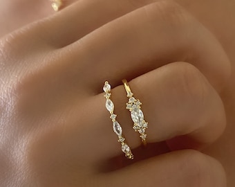 May include: Two gold rings with clear, sparkling stones. The rings are stacked on a finger. The ring on top has a larger, oval-shaped stone surrounded by smaller stones. The ring on the bottom has a row of smaller, round stones.