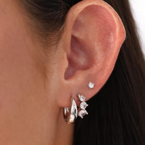 May include: An ear with multiple earrings. A silver hoop earring is in the lobe, with a cluster of three clear stones above it. A small stud earring is visible above the cluster. The earrings are set against the skin.