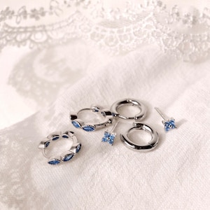 May include: A selection of silver earrings featuring blue gemstones. The jewellery set includes hoop earrings, stud earrings, and small huggie earrings. The earrings are presented on a white, textured fabric.