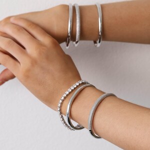May include: Several silver bracelets are displayed on wrists. The bracelets include a solid, polished band, a band with small, sparkling stones, and a textured band. The jewellery is set against a plain white background.
