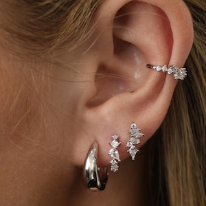 May include: A silver-toned ear cuff with clear stones, a smooth silver hoop earring, and two silver stud earrings featuring clusters of clear, multi-shaped stones. The jewelry is arranged along the earlobe and upper cartilage for a layered look.