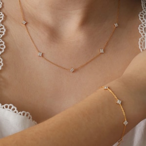 May include: A gold-colored necklace and bracelet set. Both pieces feature small, four-petal flower designs with clear, sparkling accents. The necklace is around the neck, and the bracelet is on the wrist.