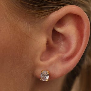 May include: A gold stud earring featuring a single oval-shaped clear gemstone held in place by four prongs. The earring is positioned on the earlobe, showcasing the reflective facets of the stone and the polished gold metal setting.