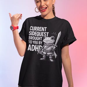 May include: Black t-shirt with the text "CURRENT SIDEQUEST BROUGHT TO YOU BY ADHD." The shirt features a cartoon frog character holding a sword. The frog is wearing a cape and armor. The background is a gradient of pink and purple.