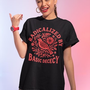 Könnte beinhalten: Schwarzes T-Shirt mit roter Grafik, die einen Vogel und Blumen sowie den Text "RADICALIZED BY BASIC DECENCY" zeigt. Das T-Shirt hat einen Rundhalsausschnitt und kurze Ärmel.