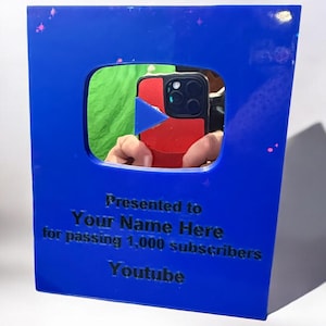 May include: A blue acrylic YouTube play button award. The award has a square shape with a cutout in the center. The text on the award reads "Presented to Your Name Here for passing 1,000 subscribers Youtube."