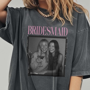 May include: Dark grey oversized t-shirt with the word "BRIDESMAID" in pink. The shirt features a black and white photo of two women laughing. The shirt is made of a soft material.
