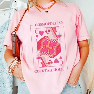 May include: Pink t-shirt with a graphic of a queen of hearts playing card with the text "COSMOPOLITAN COCKTAIL HOUR".