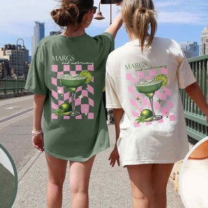 May include: Two people wearing oversized t-shirts. The shirts feature a margarita glass design with the text "MARGS and matrimony" on the back. One shirt is moss green, the other is ivory. The shirts also have the text "SAVE WATER DRINK MARGS" on the front.