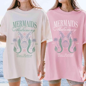 May include: Two oversized t-shirts with a mermaid and seashell graphic. The text on the shirts reads "Mermaids and Matrimony" and "Fiona's Bachelorette Rosemary Beach, FL". The shirts are in ivory and blossom colors.