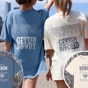 May include: Two women wearing blue and white t-shirts with the text "Gettin' Rowdy" and "Gettin' Hitched" on the back. The shirts also feature a design of a string of diamonds. The women are standing on a bridge with a view of the city in the background.
