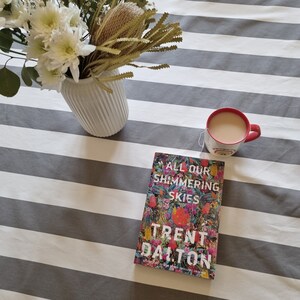 May include: A book titled "All Our Shimmering Skies" by Trent Dalton, with a colorful floral design on the cover, is lying on a striped gray and white tablecloth. A red mug with tea is next to the book.
