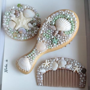 Può includere: Una spazzola per capelli in legno, uno specchio rotondo e un pettine decorati con perle bianche e verde chiaro, varie conchiglie e piccole stelle marine. Gli oggetti sono disposti su una superficie bianca con il testo "Halee.ie" stampato a sinistra.
