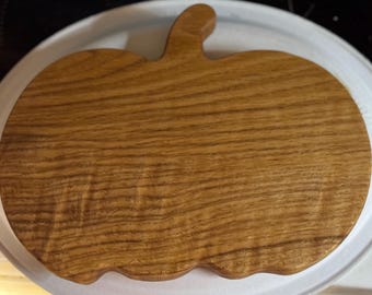 May include: A wooden pumpkin-shaped cutting board with a stem. The board is made of light brown wood with a visible grain pattern. The board is on a white plate.