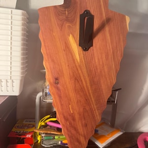 May include: A wooden arrowhead-shaped plaque with a natural reddish-brown finish. It features a dark metal bracket for hanging. The wood grain is visible, and the edges are uneven, giving it a rustic appearance.
