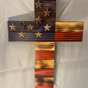 May include: A wooden cross with the American flag design. The cross is made of multiple pieces of wood, each painted in red, white, and blue. The stars are painted in gold.