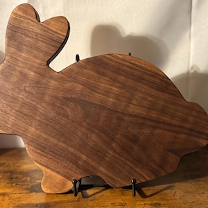 May include: A brown wooden cutting board shaped like a rabbit, with a dark wood grain and a natural finish. The board is on a stand with two black metal legs.