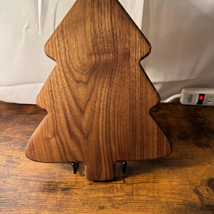 May include: A wooden Christmas tree-shaped cutting board. The board is made of a dark brown wood with visible grain patterns. It has a flat surface and is supported by two small, black metal stands.