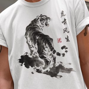 May include: White t-shirt featuring a black and gray tiger illustration with Chinese characters. The tiger is depicted in a traditional ink wash painting style. The shirt has rolled-up sleeves.