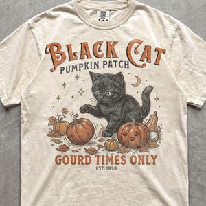 Könnte beinhalten: Ein elfenbeinfarbenes Kurzarm-T-Shirt mit einer Grafik einer schwarzen Katze in einem Kürbisfeld. Der Text lautet BLACK CAT PUMPKIN PATCH, GOURD TIMES ONLY und EST. 1898. Das Design umfasst orangefarbene Kürbisse, Herbstblätter, Sterne und eine Mondsichel.