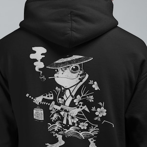 Könnte beinhalten: Schwarzer Kapuzenpullover mit einer weißen Illustration einer Froschfigur im Kimono, die eine Pfeife raucht und ein Schwert und eine Laterne hält. Der Frosch trägt einen japanischen Hut. Das Design umfasst Blumen- und Wellenmuster.