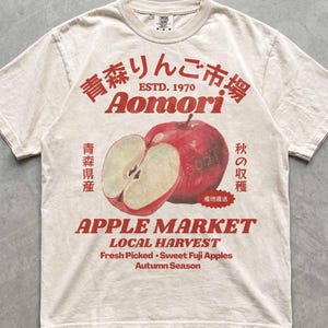 Könnte beinhalten: Ein elfenbeinfarbenes Kurzarm-T-Shirt mit einer roten Grafik eines ganzen und eines aufgeschnittenen Apfels. Der Text lautet Aomori, Apple Market, Local Harvest, Fresh Picked, Sweet Fuji Apples, Autumn Season, Estd. 1970 sowie japanische Schriftzeichen.